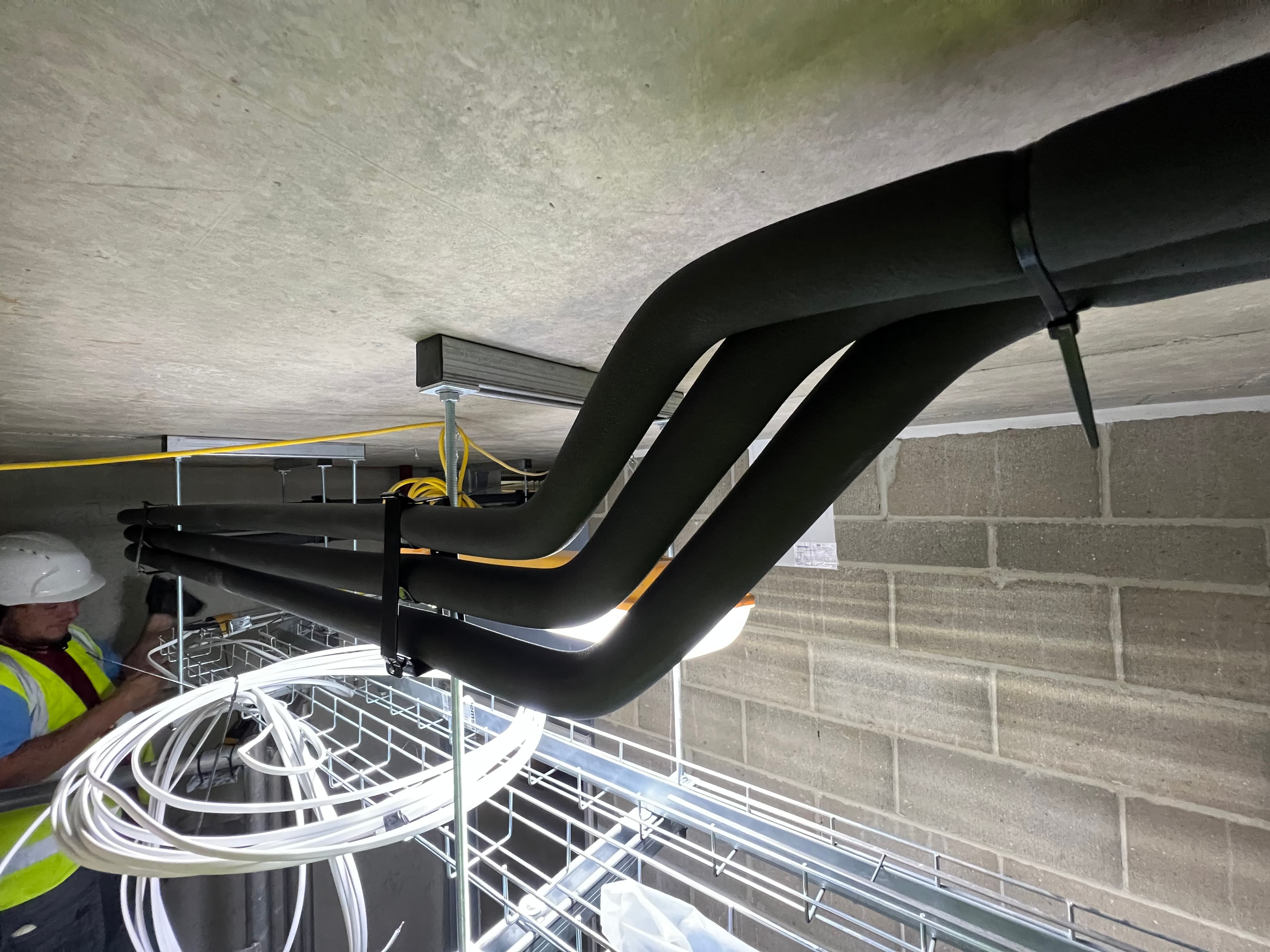 Construction worker installs black insulated pipes and white cables along a concrete ceiling.